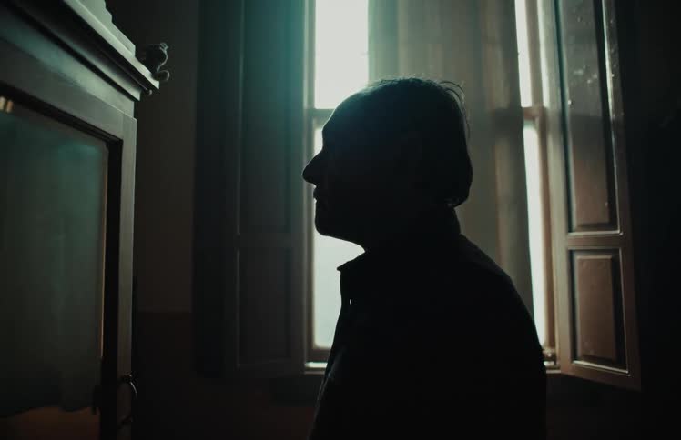 A contemplative man in silhouette stands in a dark, vintage room, illuminated by the light from a window