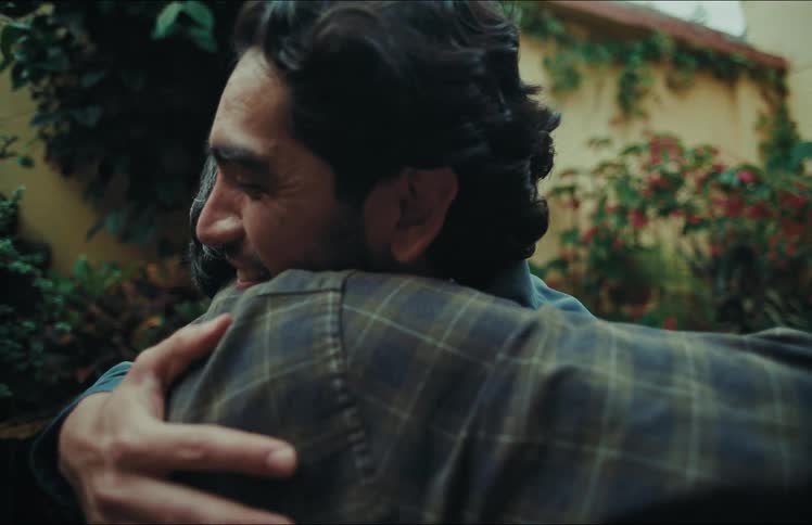 Two men share an emotional and heartfelt embrace in a garden courtyard