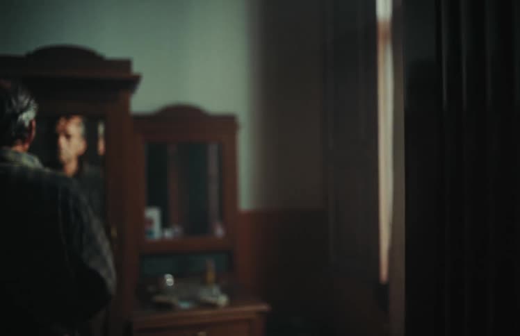 A pensive older man with gray hair gazes at his reflection in a mirror on a vintage wardrobe in a dark, moody room