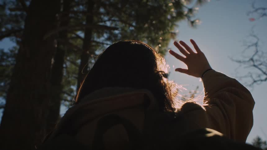 A woman with a backpack looks up and shields her eyes from the sun in the forest, creating a beautiful lens flare