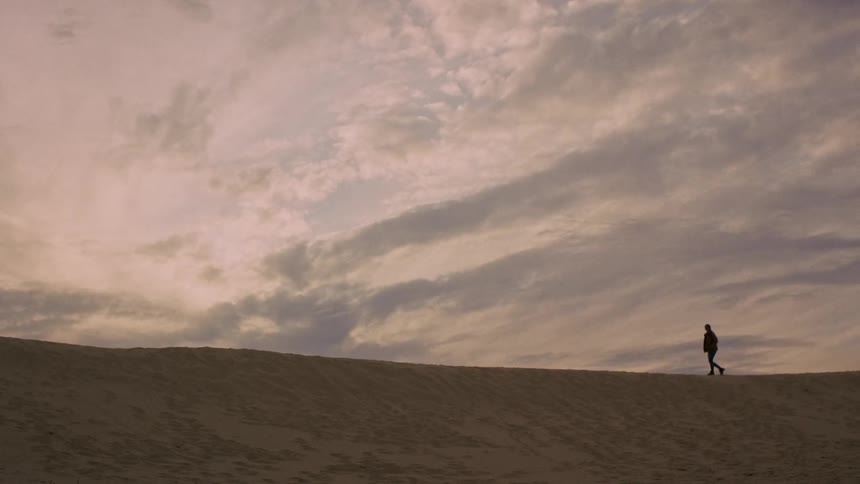 A person in silhouette walks across a sand dune horizon before stopping to take in the view