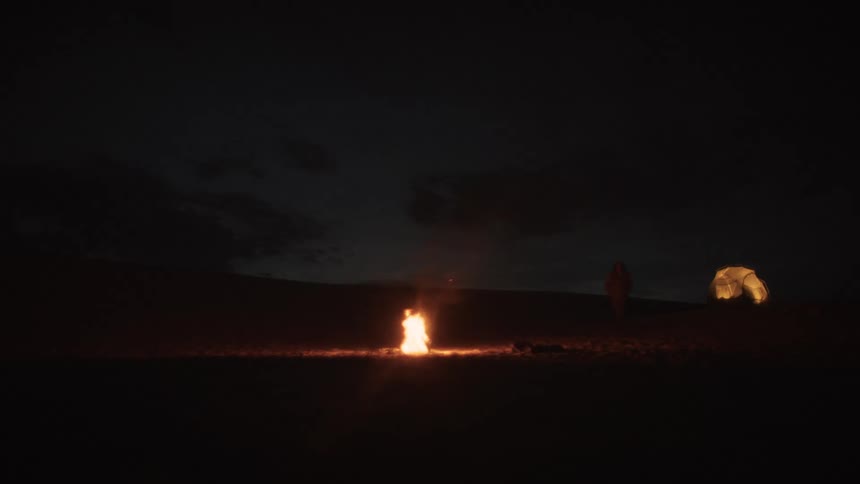 A solitary person walks from an illuminated tent to a warm campfire in a vast, dark landscape at night