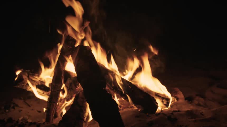 A cozy campfire crackles in slow motion on a sandy beach at night