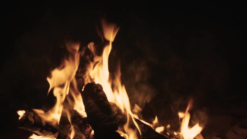 A close-up view of a crackling campfire at night, with bright orange flames licking logs and sparks rising into the darkness