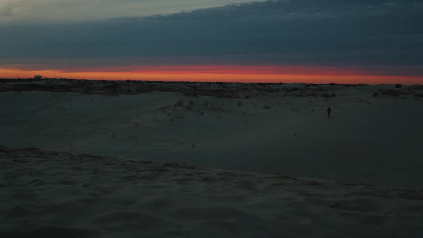 A lone figure walks across a vast desert landscape against a dramatic and colorful sunset