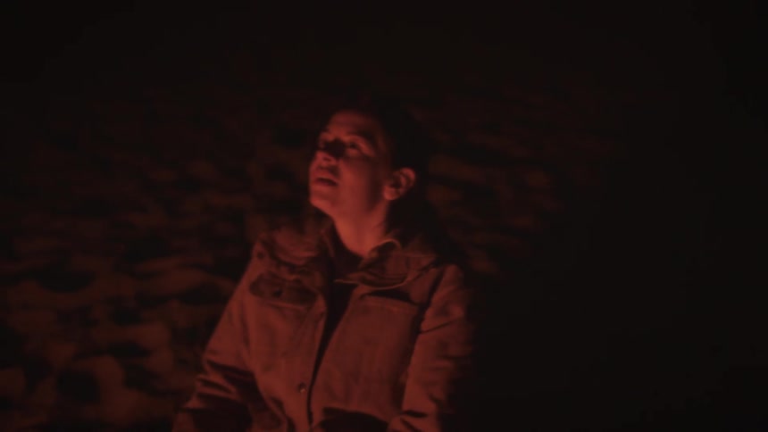 A woman's face, bathed in the warm, red light of a campfire, looks up peacefully as the camera slowly pulls out, leaving her in darkness