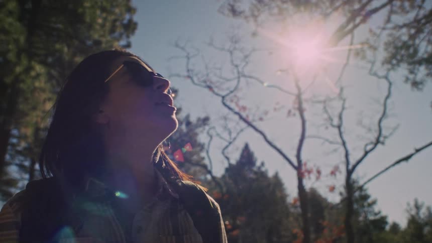 A smiling woman in sunglasses soaks in the warm sunlight while standing among tall forest trees in slow motion