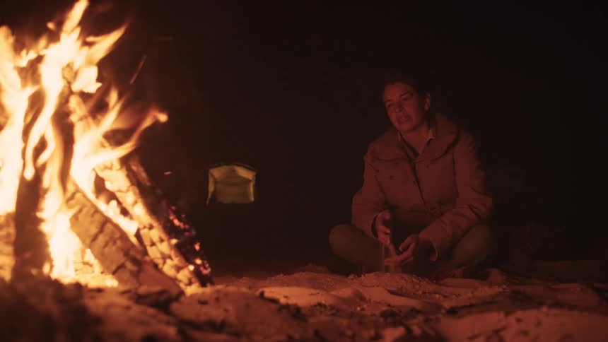A peaceful woman sits alone by a roaring campfire on the sand at night, enjoying a moment of solitude while camping