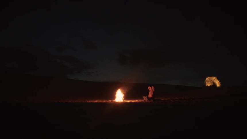 A solitary figure in robes prays by a campfire at night in the vast desert wilderness