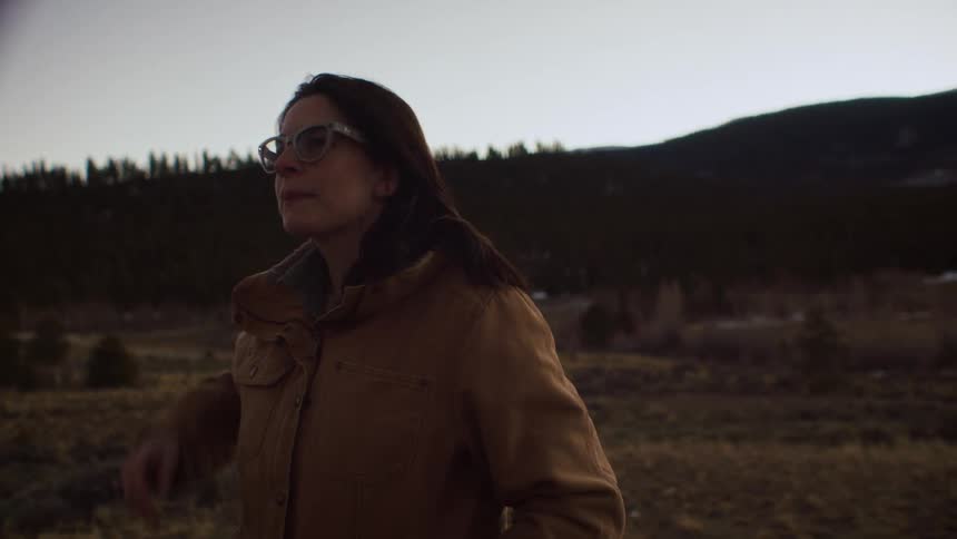 A thoughtful woman wearing glasses and a warm jacket takes in the quiet beauty of the mountain landscape at dusk in slow motion