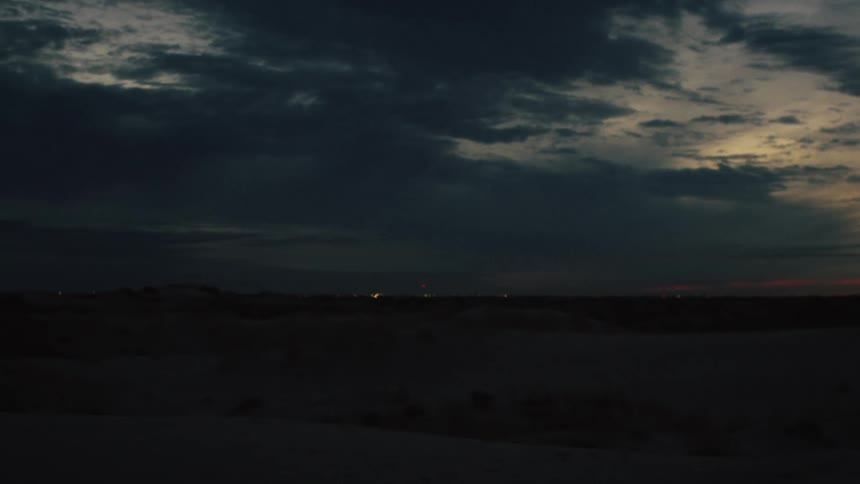 Distant city lights twinkle on the horizon of a dark, vast desert landscape under a cloudy twilight sky