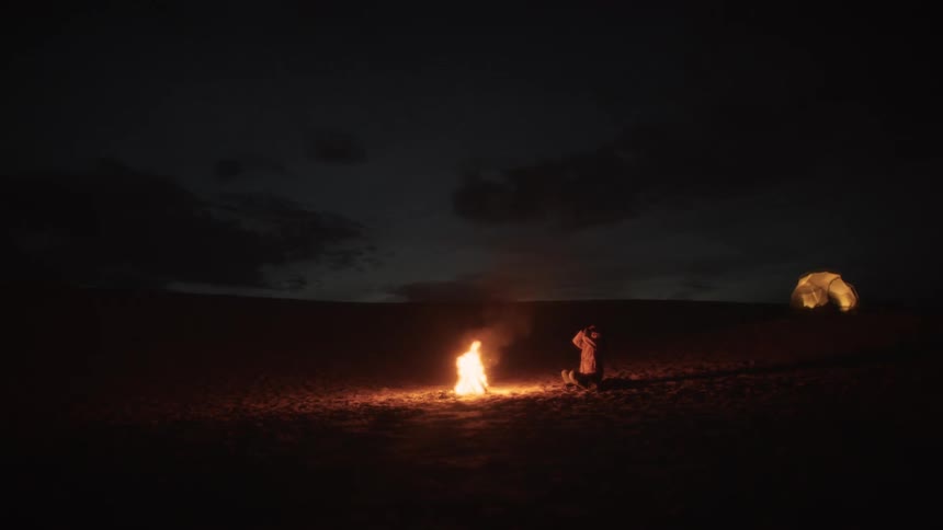A serene night in the desert as a traveler warms themselves by a campfire near their glowing tent