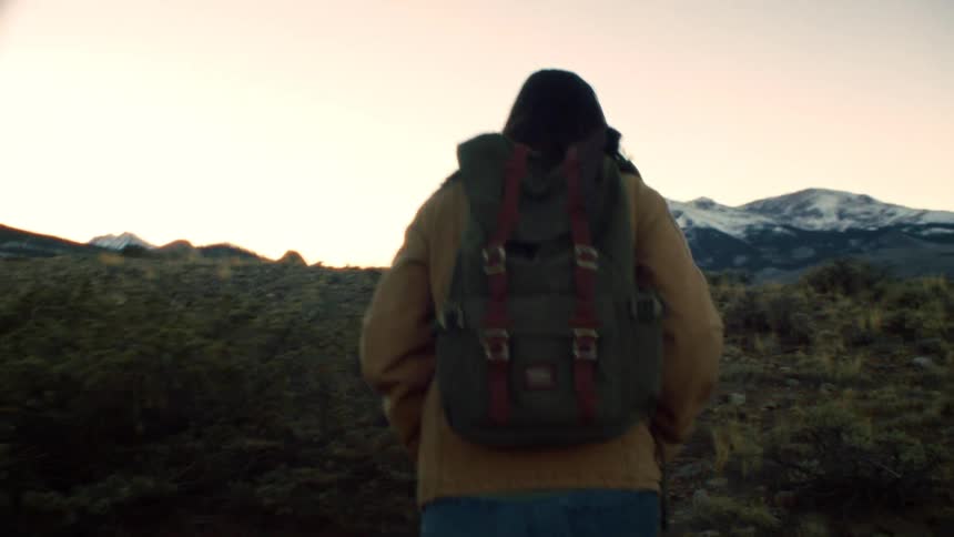 Slow-motion tracking shot from behind a hiker with a backpack walking through a mountain landscape at sunset