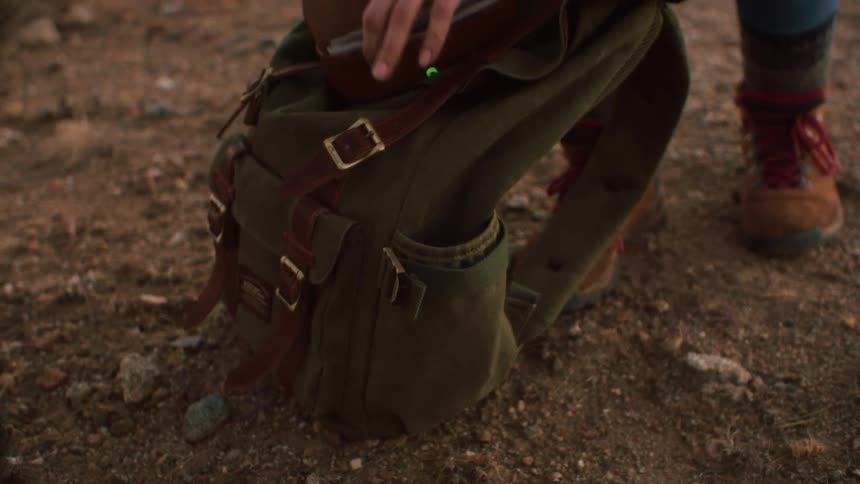 A hiker stops on a trail to pull a glasses case from their green canvas backpack