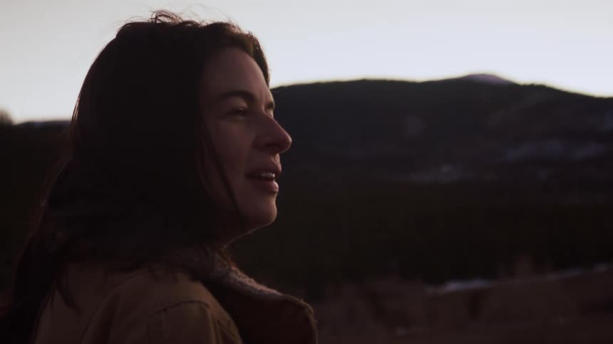A woman in profile smiles peacefully as she looks out over a beautiful mountain landscape at sunset in slow motion