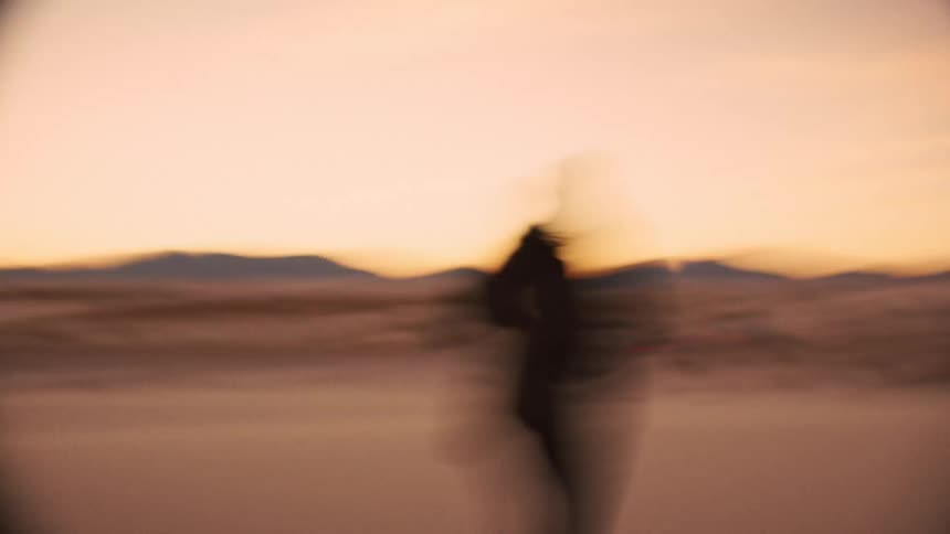 Shaky and blurry footage captures the silhouette of a person wandering through a vast desert landscape at sunset