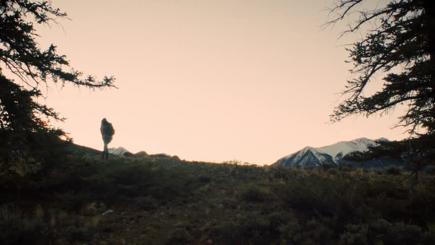 A silhouetted hiker with a backpack walks along a ridgeline at sunrise with a view of snow-capped mountains