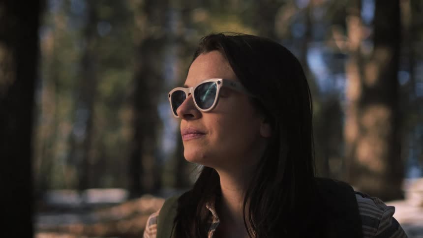 A slow-motion close-up shot of a thoughtful woman wearing sunglasses in a sunlit forest
