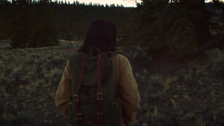 A woman with a backpack hikes through a rugged wilderness landscape towards snow-capped mountains at dusk