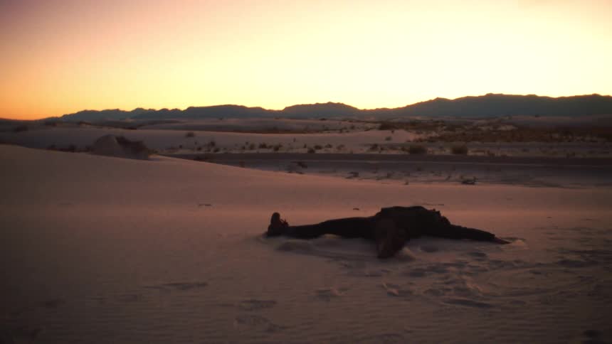 A person lies on a desert sand dune making a sand angel at sunset with mountains in the background