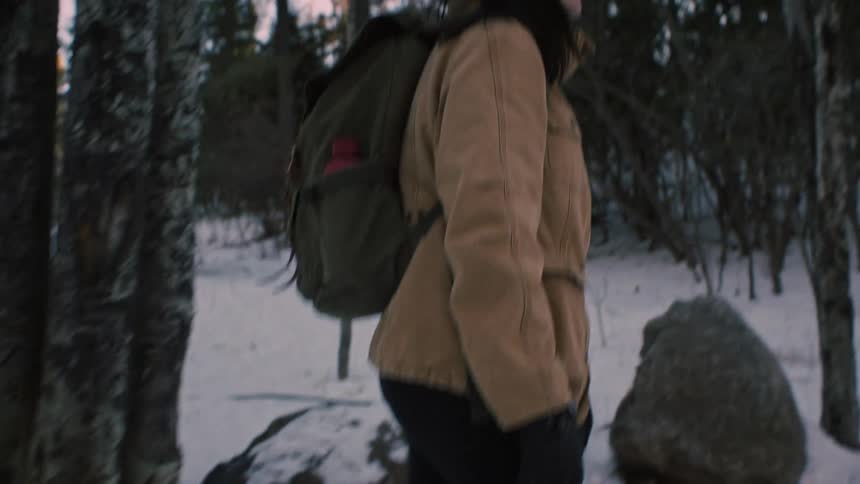 A woman wearing hiking boots and a backpack walks through a snowy winter forest and stops to admire the tall trees