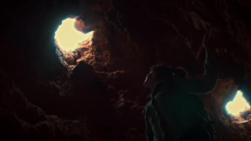 A silhouetted woman inside a dark cave looks up towards the sunlight pouring in from holes in the ceiling