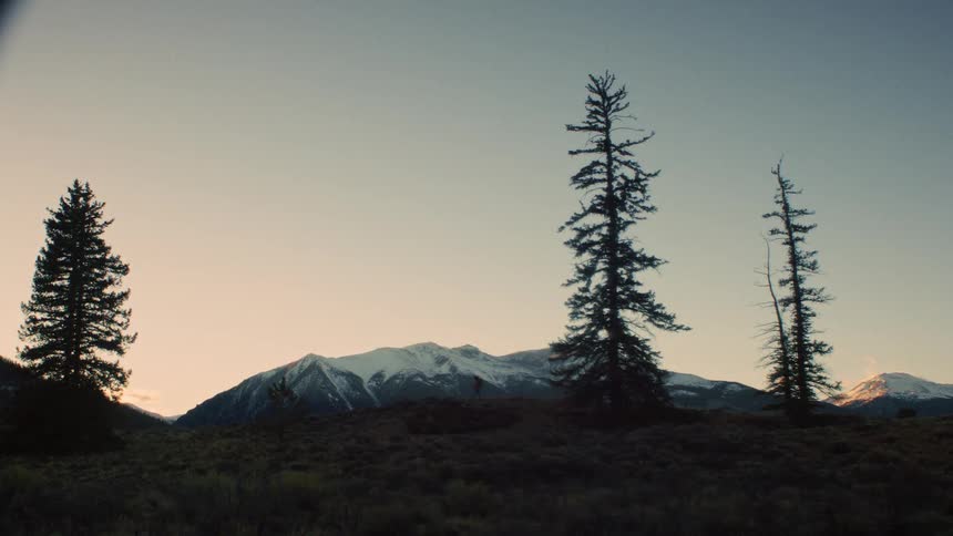 A lone hiker is silhouetted against the rising sun as they walk along a mountain ridge with snow-capped peaks in the distance