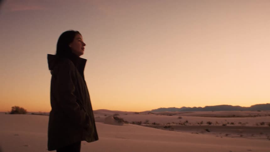 A contemplative woman in a jacket watches the golden sunset over the white sand dunes
