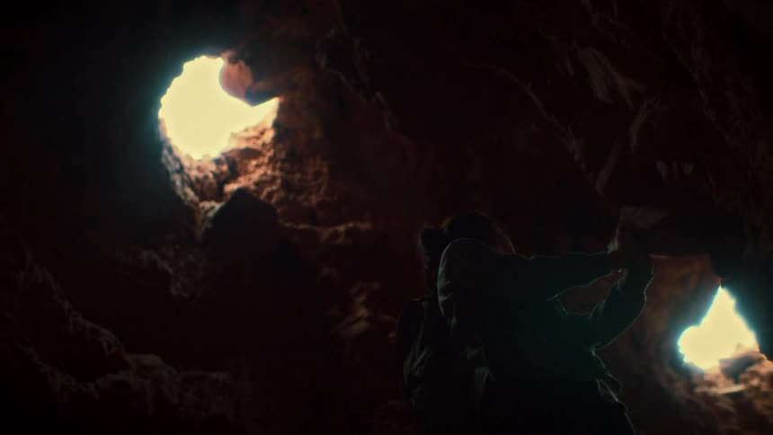From a low angle, two explorers are silhouetted inside a dark cave as they look up towards an opening to the sky