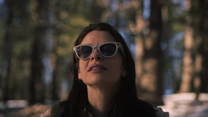 A slow-motion close-up of a woman in sunglasses looking up with a gentle smile as she takes in the scenery of a sunny, snowy forest