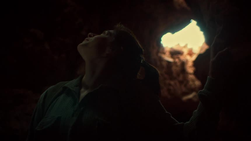 A curious woman stands in the darkness of a cave, looking up with hope at the light pouring through an opening in the rock ceiling