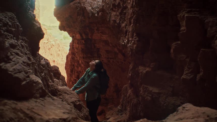 A female explorer with a backpack stands inside a dark, narrow cave and looks up in awe at the light pouring in from above