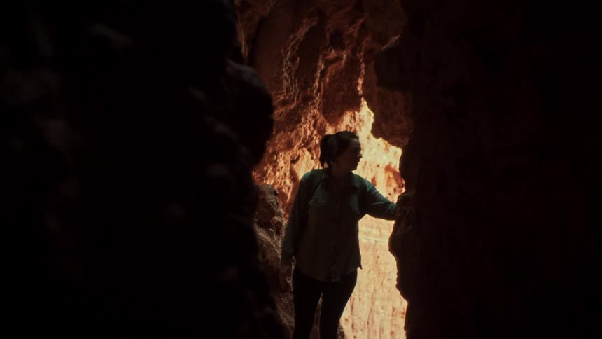 A curious woman stands silhouetted inside a dark cave, looking up in awe at the sunlit red rock formations