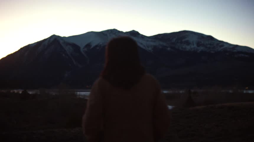 A mysterious woman turns to look back over her shoulder in slow motion against a backdrop of snowy mountains at twilight
