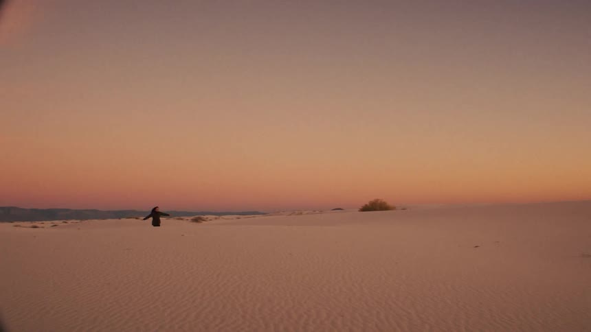 A solitary person in a flowing robe wanders through a serene desert of white sand under a colorful sunset sky