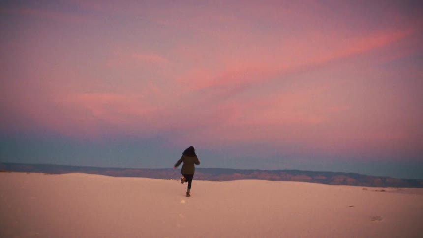 A woman joyfully runs away from the camera across vast white sand dunes during a beautiful pink sunset