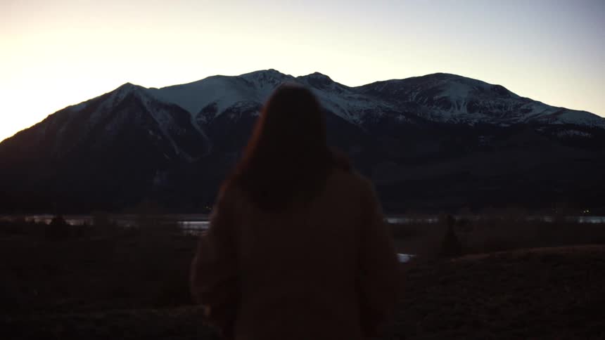 A woman stands silhouetted against the fading light, looking out at a majestic snow-capped mountain range at sunset
