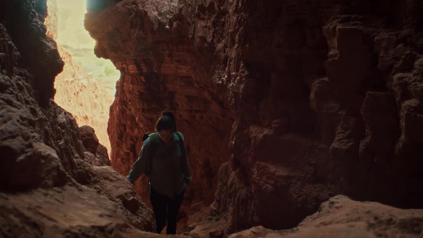An adventurous woman with a backpack carefully explores the inside of a dark, narrow red rock slot canyon