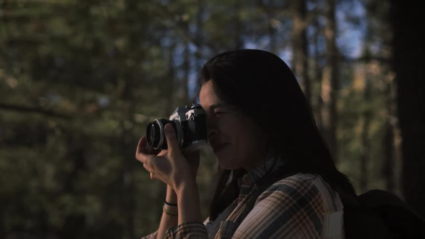 A smiling young woman with a backpack happily takes pictures with her vintage camera while hiking in a beautiful sunlit forest