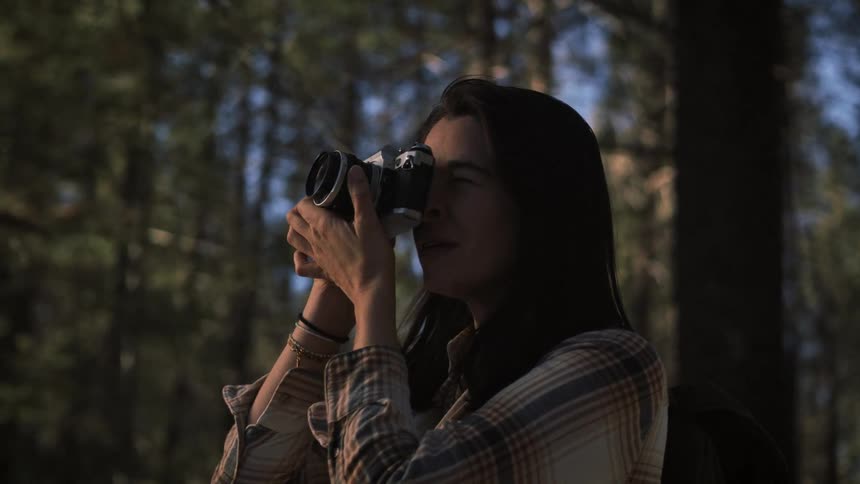 A female hiker peacefully takes a photograph with her vintage film camera while exploring a beautiful sunlit forest