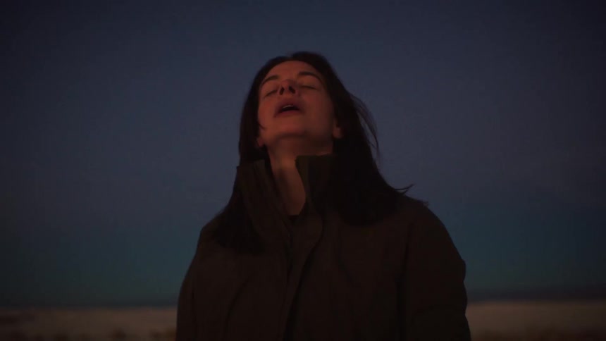 A tranquil woman with her eyes closed takes a deep, peaceful breath outdoors against the dark twilight sky