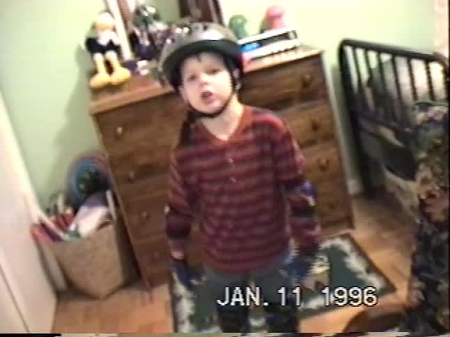 A nostalgic 1996 home video shows a young boy fully geared up with a helmet, elbow pads, and gloves in his bedroom