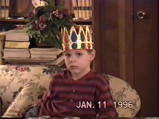A sleepy young boy wearing a colorful crown yawns while sitting on a couch in a vintage 1996 home video