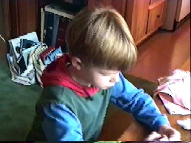 A young boy intently colors an Easter egg with crayons at a dining table in this nostalgic home video