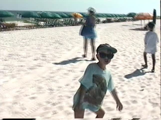 Vintage home video footage of a mother and her young son walking along a sunny, white sand beach