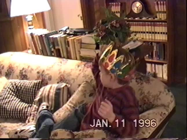 A young boy wearing a paper crown sits on a floral couch in a cozy living room in a vintage 1996 home video