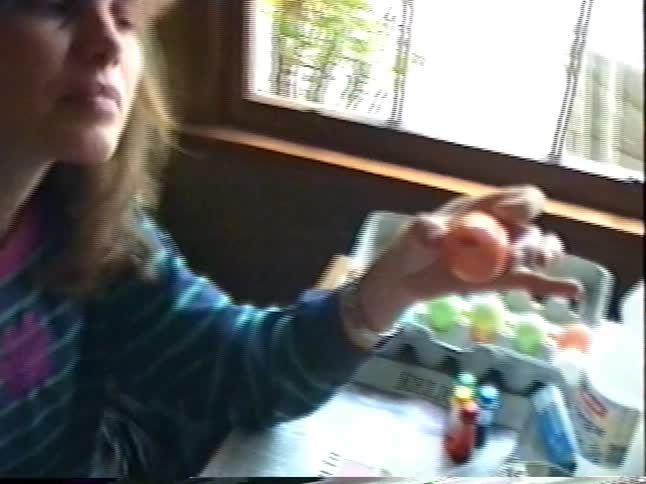 A mother shows off a dyed orange Easter egg while sitting at a dining table with her child in this 1990s home video