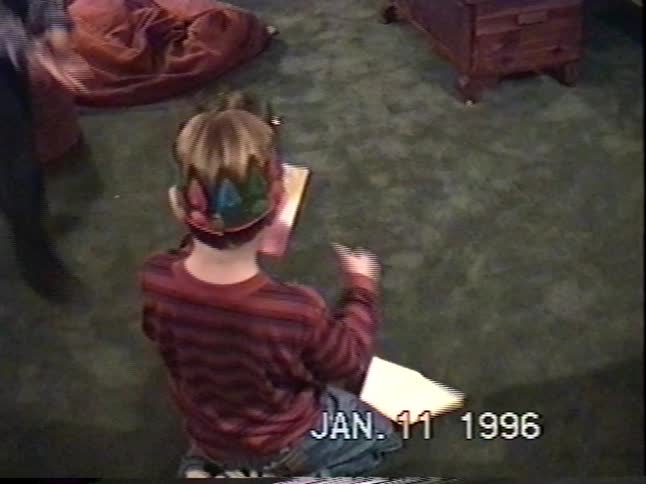Young boy wearing a colorful crown opens a birthday card while sitting on the living room floor in a 1996 home video