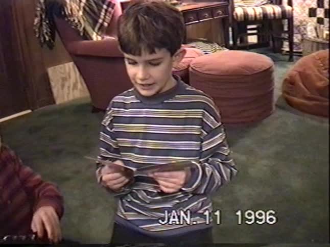 A young boy reads a birthday card aloud while his sibling watches during a family celebration in this vintage 1996 home video