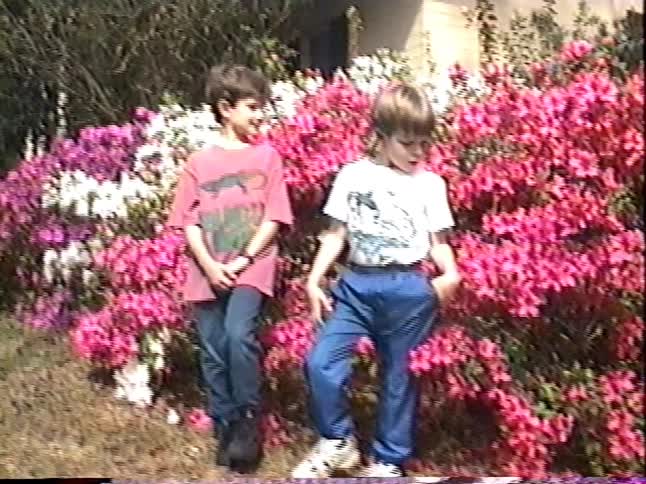 Two young brothers stand in front of a vibrant bush of pink flowers for a picture in this nostalgic home video from the 1990s